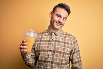 Young handsome man drinking healthy orange juice using straw over yellow background with a happy face standing and smiling with a confident smile showing teeth