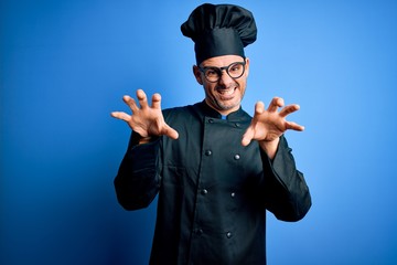 Young handsome chef man wearing cooker uniform and hat over isolated blue background smiling funny...