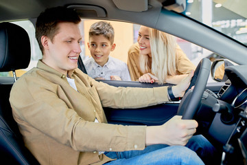 Happy young caucasian car owner in dealership,handsome young man sitting at front seat of the car, likes design of auto, wife and son waiting for his reaction on car, ask about feelings