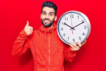 Young handsome man with beard holding big clock smiling happy and positive, thumb up doing excellent and approval sign