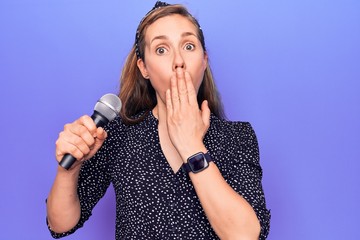 Young beautiful blonde woman holding microphone singing over purple background covering mouth with...