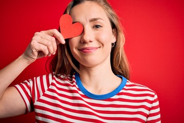 Young beautiful blonde woman holding paper heart standing over isolated red background with a happy face standing and smiling with a confident smile showing teeth