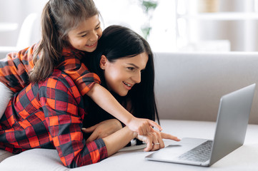 Fototapeta premium Cheerful and smiling caucasian mom and her cute daughter are communicate by video conference with grandparents by laptop while lying at home on the couch