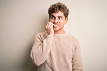 Young blond handsome man with curly hair wearing casual sweater over white background looking stressed and nervous with hands on mouth biting nails. Anxiety problem.