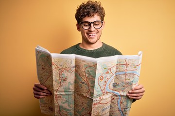 Young blond tourist man on vacation with curly hair holding city map over yellow background with a happy face standing and smiling with a confident smile showing teeth