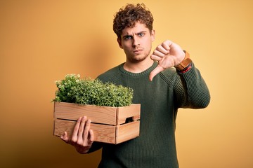 Young blond handsome man with curly hair holding wooden box with plants with angry face, negative...