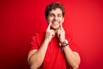 Young blond handsome man with curly hair wearing casual t-shirt over red background Smiling with open mouth, fingers pointing and forcing cheerful smile