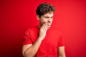 Young blond handsome man with curly hair wearing casual t-shirt over red background touching mouth with hand with painful expression because of toothache or dental illness on teeth. Dentist
