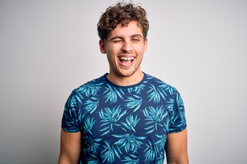 Young blond handsome man on vacation with curly hair wearing casual summer t-shirt winking looking...
