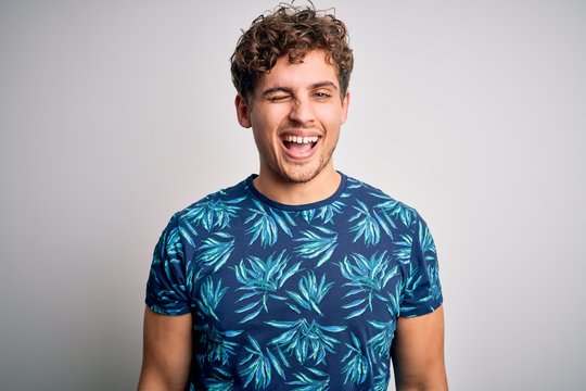 Young blond handsome man on vacation with curly hair wearing casual summer t-shirt winking looking at the camera with sexy expression, cheerful and happy face.