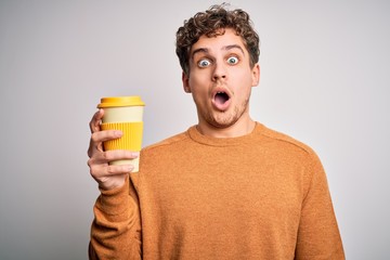 Young blond man with curly hair drinking cup of coffee standing over white background scared in shock with a surprise face, afraid and excited with fear expression