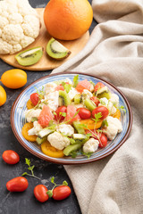 Vegetarian salad of cauliflower cabbage, kiwi, tomatoes, microgreen sprouts on black concrete background. Side view, close up.