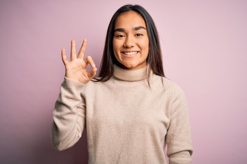 Young beautiful asian woman wearing casual turtleneck sweater over pink background smiling positive doing ok sign with hand and fingers. Successful expression.