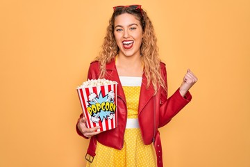 Beautiful blonde woman with blue eyes eating salty popcorn snack over yellow background screaming proud and celebrating victory and success very excited, cheering emotion
