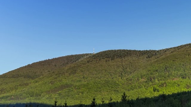 Zoom in hyperlapse tower timelapse of shadows and clouds moving on Radhost Hill overlooking a tower in a dense forest with moving shadows reflecting the sun from clouds Beskydy Park area.