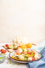 Vegetarian salad of cauliflower cabbage, kiwi, tomatoes, microgreen sprouts on gray wooden background. Side view, selective focus, copy space.