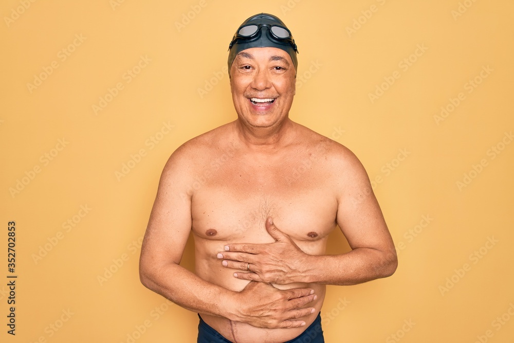 Poster Middle age senior grey-haired swimmer man wearing swimsuit, cap and goggles smiling and laughing hard out loud because funny crazy joke with hands on body.