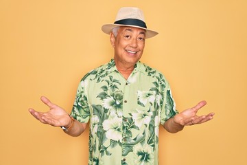 Middle age senior grey-haired man wearing summer hat and floral shirt on beach vacation clueless and confused expression with arms and hands raised. Doubt concept.