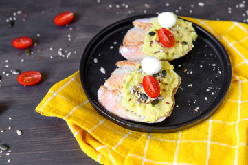 Two sandwiches with avocado and cherry tomatoes and mozzarella with seeds of flax, sesame, pumpkin and half-sunflower in a black ceramic plate on a wooden table with a yellow napkin.