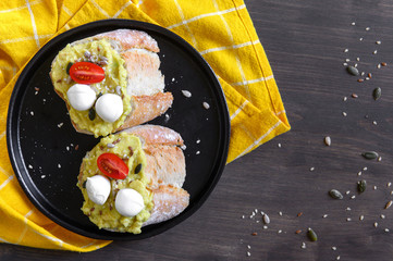 Two sandwiches with avocado and cherry tomatoes and mozzarella with seeds of flax, sesame, pumpkin and half-sunflower in a black ceramic plate on a wooden table with a yellow napkin. Flat lay.