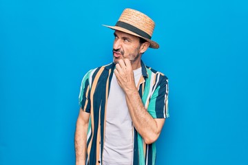 Middle age handsome tourist man on vacation wearing shirt and hat over blue background thinking concentrated about doubt with finger on chin and looking up wondering