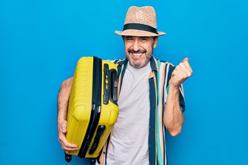 Middle age handsome tourist man on vacation holding cabin bag over isolated blue background screaming proud, celebrating victory and success very excited with raised arm