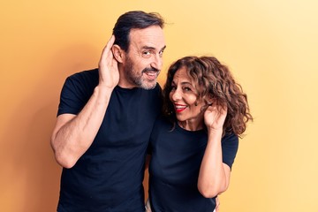 Middle age beautiful couple wearing t-shirt standing over isolated yellow background smiling with hand over ear listening and hearing to rumor or gossip. Deafness concept.