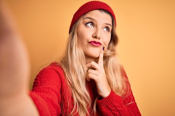 Young beautiful blonde woman making selfie by camera over isolated yellow background serious face thinking about question, very confused idea