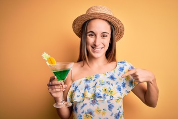 Young beautiful tourist woman on vacation wearing summer hat drinking cocktail beverage with surprise face pointing finger to himself