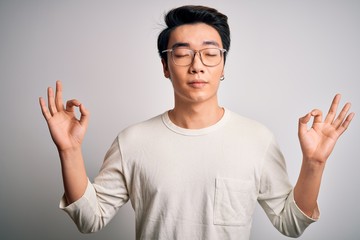 Young handsome chinese man wearing casual t-shirt and glasses over white background relax and smiling with eyes closed doing meditation gesture with fingers. Yoga concept.