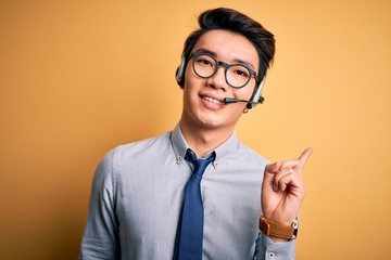 Young handsome chinese call center agent man wering glasses using headset with a big smile on face, pointing with hand and finger to the side looking at the camera.