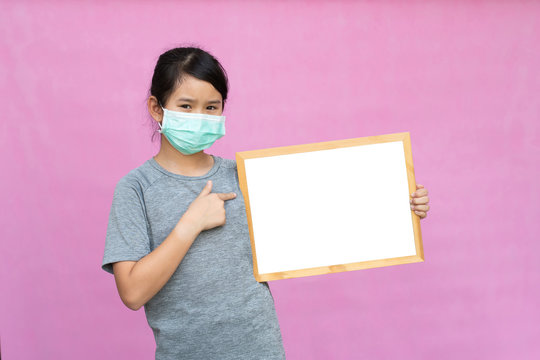 Little Asian Girl In A Protective Medical Mask Holding White Board Isolated On Pink Background. Protect From Coronavirus Or Covid-19 Epidemic.