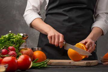 Chef cuts oranges for cooking dishes on the background of the ingredients of vegetables and fruits. Useful and proper nutrition
