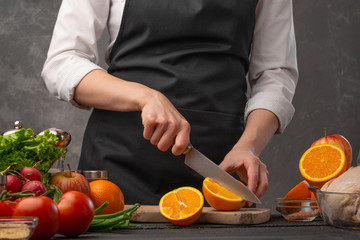 Chef cuts oranges for cooking festive chicken. Against the background of vegetables
