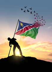 Namibia flag turn to birds while being planted by a man on a hill during sunrise. - obrazy, fototapety, plakaty