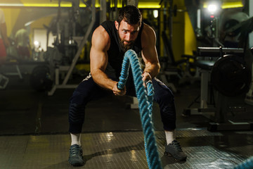 Fototapeta na wymiar Fronz view portrait of young adult caucasian man male athlete training at the dark gym working out using battle rope wearing black shirt and beard strength and endurance training