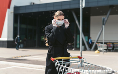 Young woman with a grocery cart on a supermarket background. Social distancing during the Coronavirus Covid-19 pandemic. Disposable mask on face and gloves on hands of a girl. Quarantine shopping