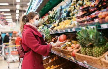Social distancing in a supermarket. A young woman in a disposable face mask buying fruits and vegetables and putting them in a grocery basket. Shopping during the Coronavirus Covid-19 epidemic 2020