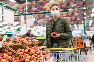Social distancing in a supermarket. A young woman in a disposable facial mask buying fruits and vegetables and putting them in a grocery basket. Shopping during the Coronavirus Covid-19 epidemic 2020