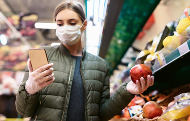 A young woman in a disposable face mask is checking a shopping list on a smartphone in a supermarket. Social distancing during the pandemic of the coronavirus Covid-19