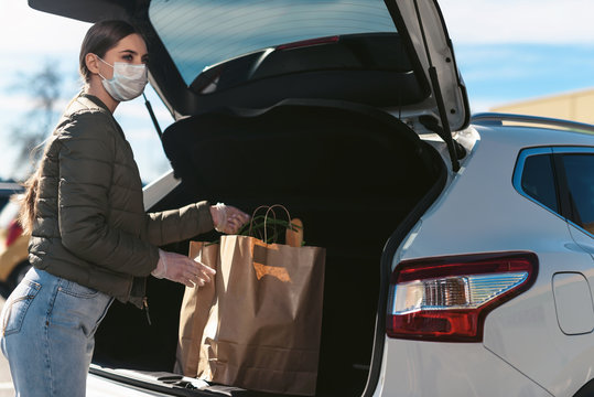 A young woman putting groceries from a supermarket in the car trunk. Social distancing: face mask, disposable gloves to prevent infection. Food shopping during coronavirus Covid-19 quarantine