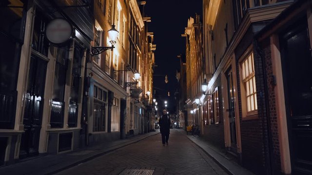 Young woman walks at empty night illuminated Amsterdam center street with closed cafes and bars, Netherlands