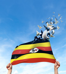 Two hands are holding a Uganda flag which morphs into birds while waving against a blue sky background. 3D Illustration.