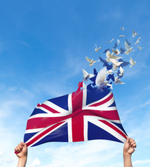 Two hands are holding a United Kingdom flag which morphs into birds while waving against a blue sky background. 3D Illustration.