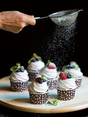 vertical photo, cream chocolate cakes, raspberry blueberry cupcake garnished with mint and icing sugar, homemade cakes on a black background, a wooden plate with tasty sweets