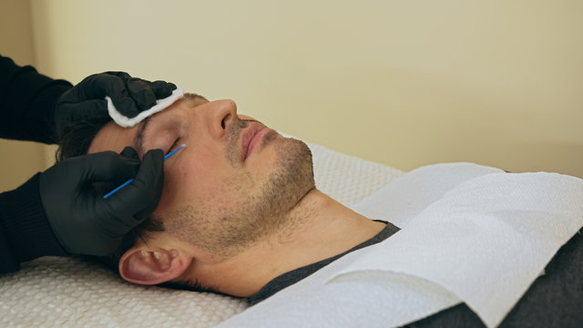 Close up of cosmetologist preparing man for the eyelash extension procedure. High quality close-up of a man laying on a couch during the esthetician lash treatment.