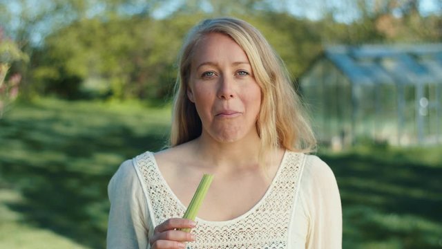 Healthy woman eats raw organic celery stalks, a greenhouse in the background