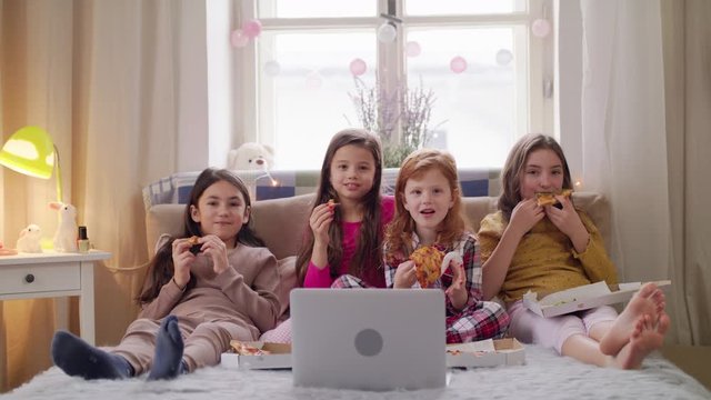Group of small girls friends watching film on bed on slumber party, eating pizza.