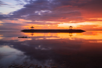 Fototapeta na wymiar Sunrise seascape. Traditional gazebos on an artificial island in the ocean. Water reflection. Bright sunlight at horizon. Couldy sky. Sanur beach, Bali, Indonesia.