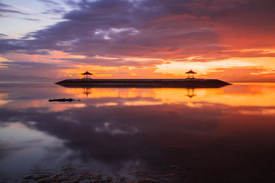 Sunrise seascape. Traditional gazebos on an artificial island in the ocean. Water reflection. Bright sunlight at horizon. Couldy sky. Sanur beach, Bali, Indonesia.